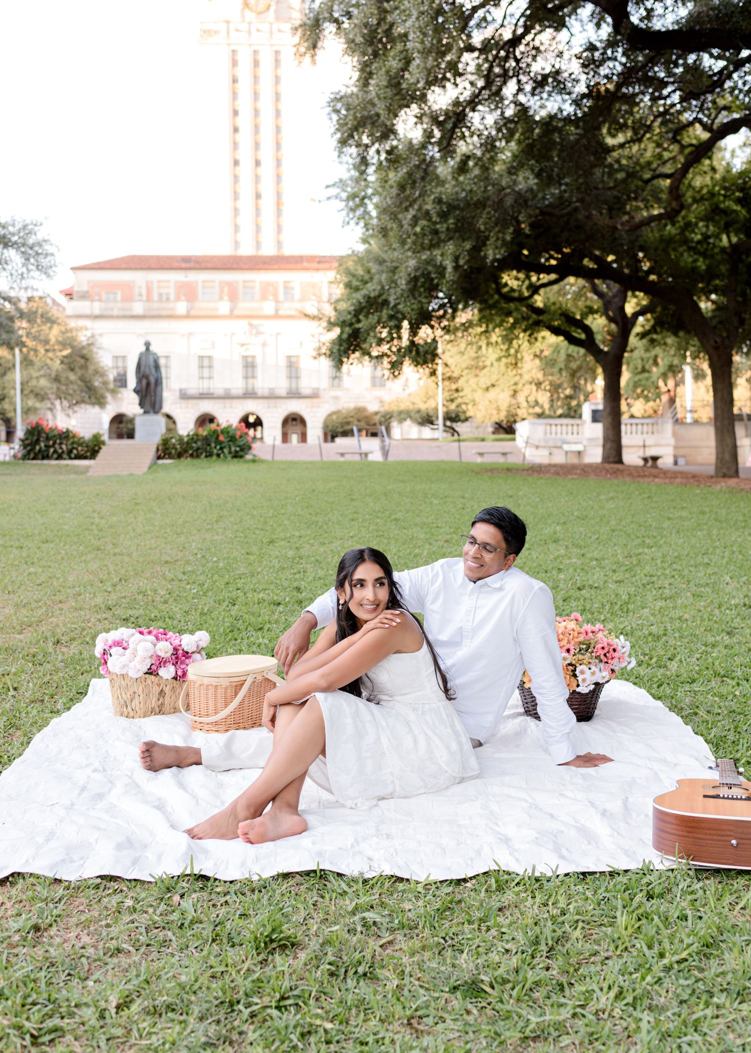 A couple enjoying a romantic picnic on a white blanket with flowers and baskets on a grassy lawn near a university building.