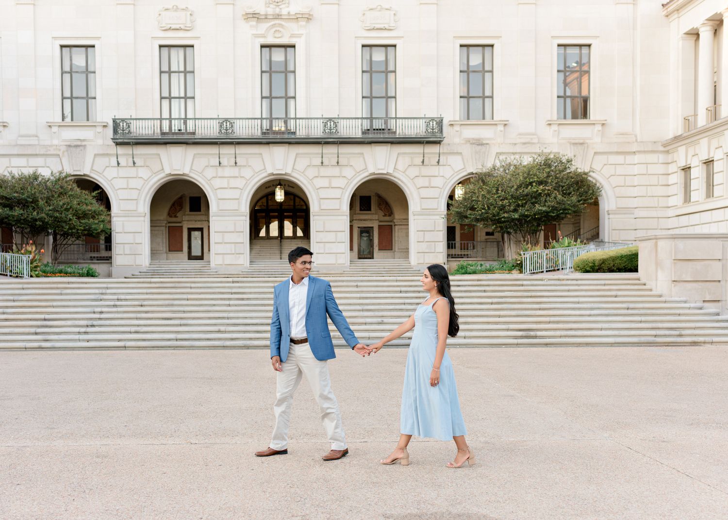 A couple in blue holds hands while walking across a grand stone building plaza with arched architecture.