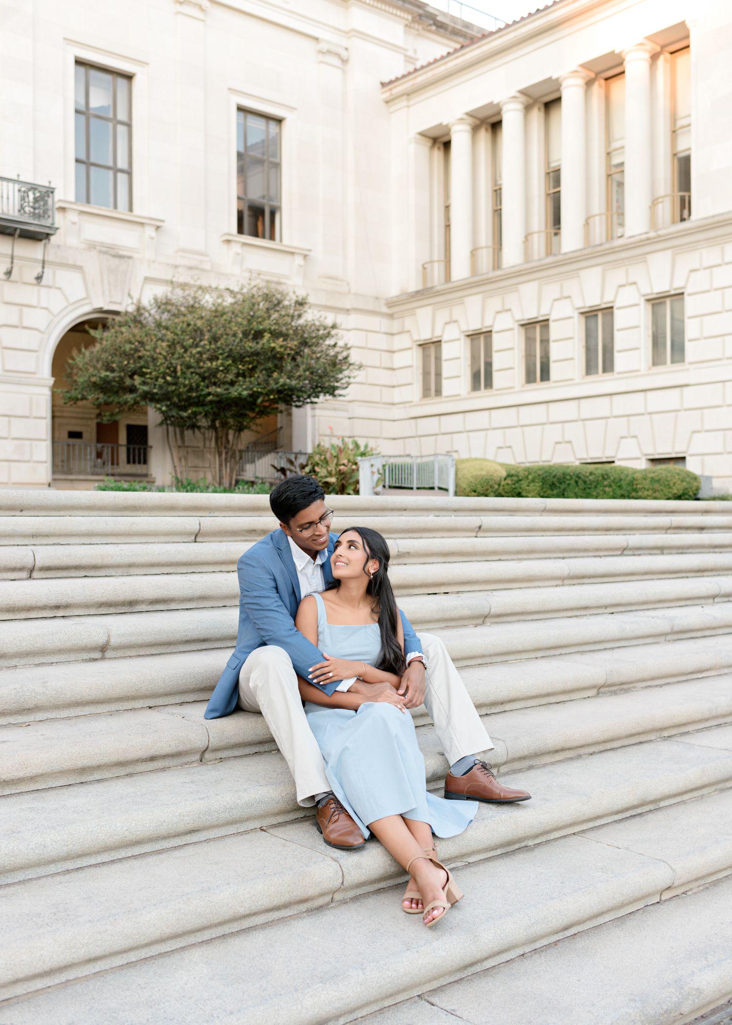 A couple shares a romantic moment while sitting on grand marble steps of a neoclassical building on a sunny day.