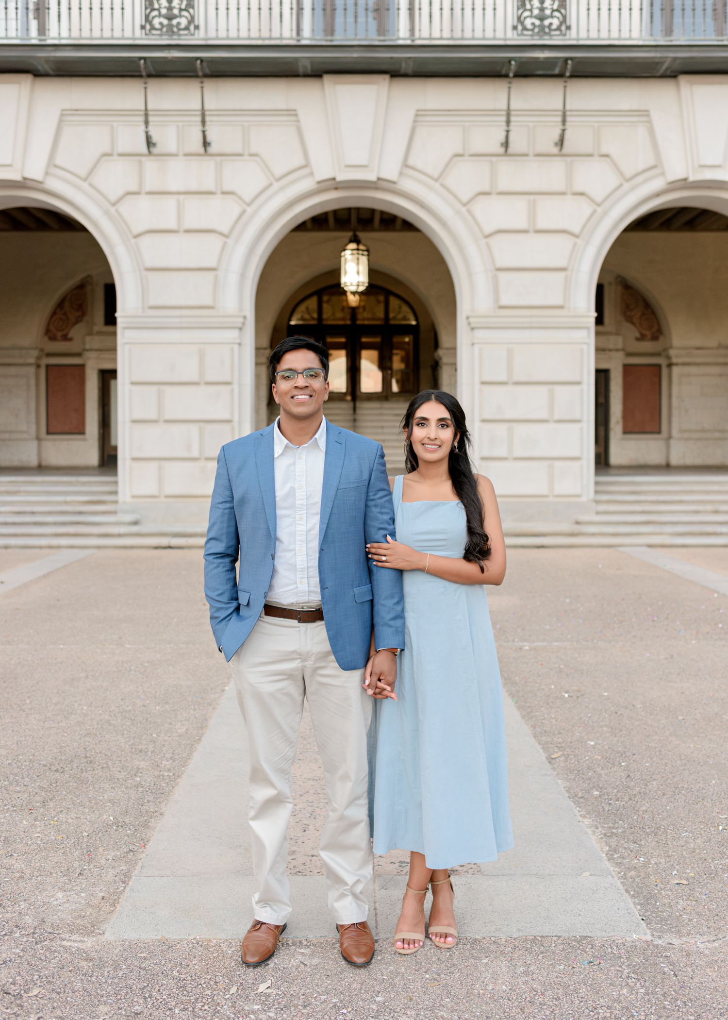 A couple stands elegantly in formal attire under white architectural arches of a grand building.