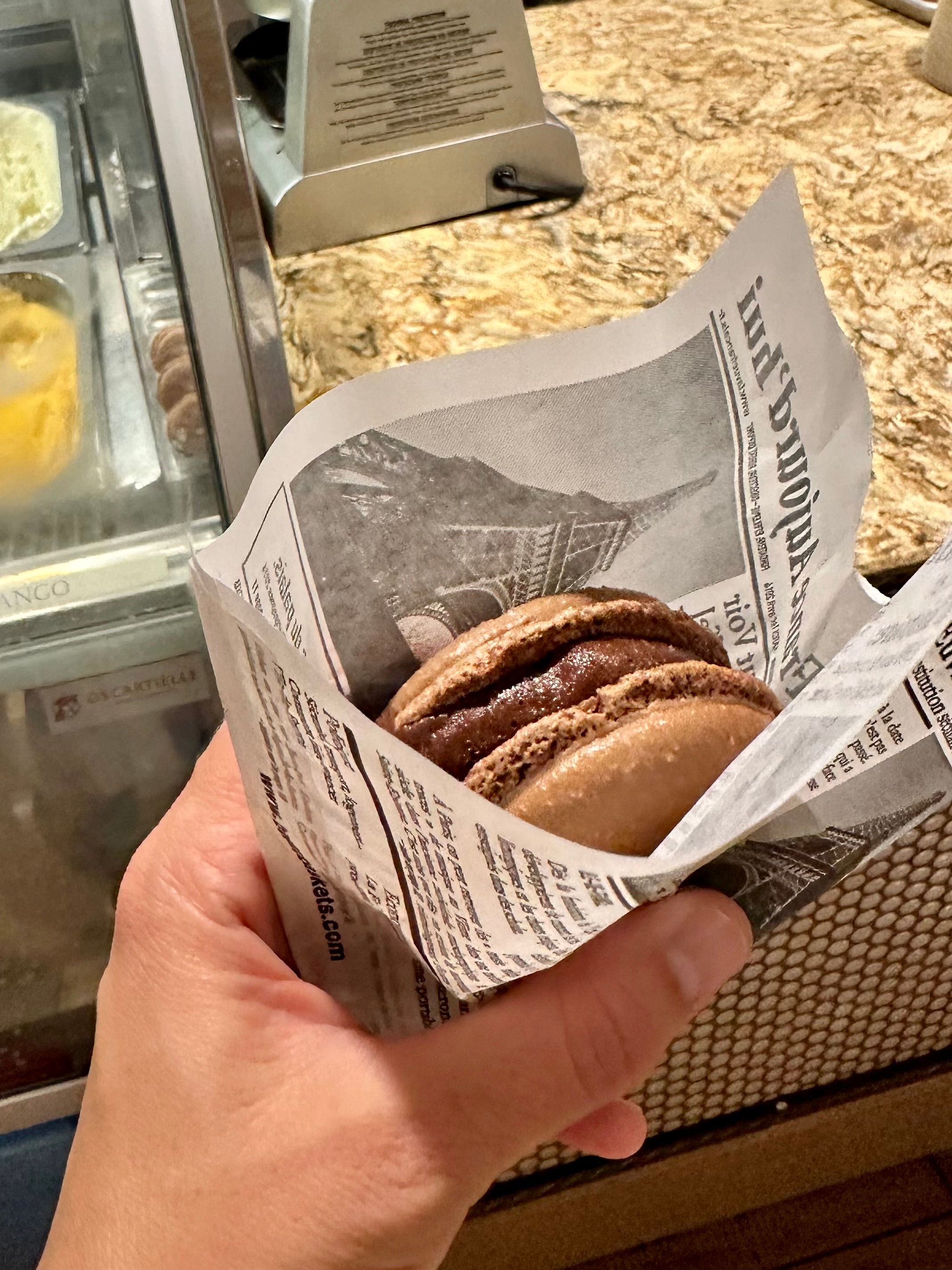 A chocolate macaron wrapped in newspaper style paper being held over a counter.