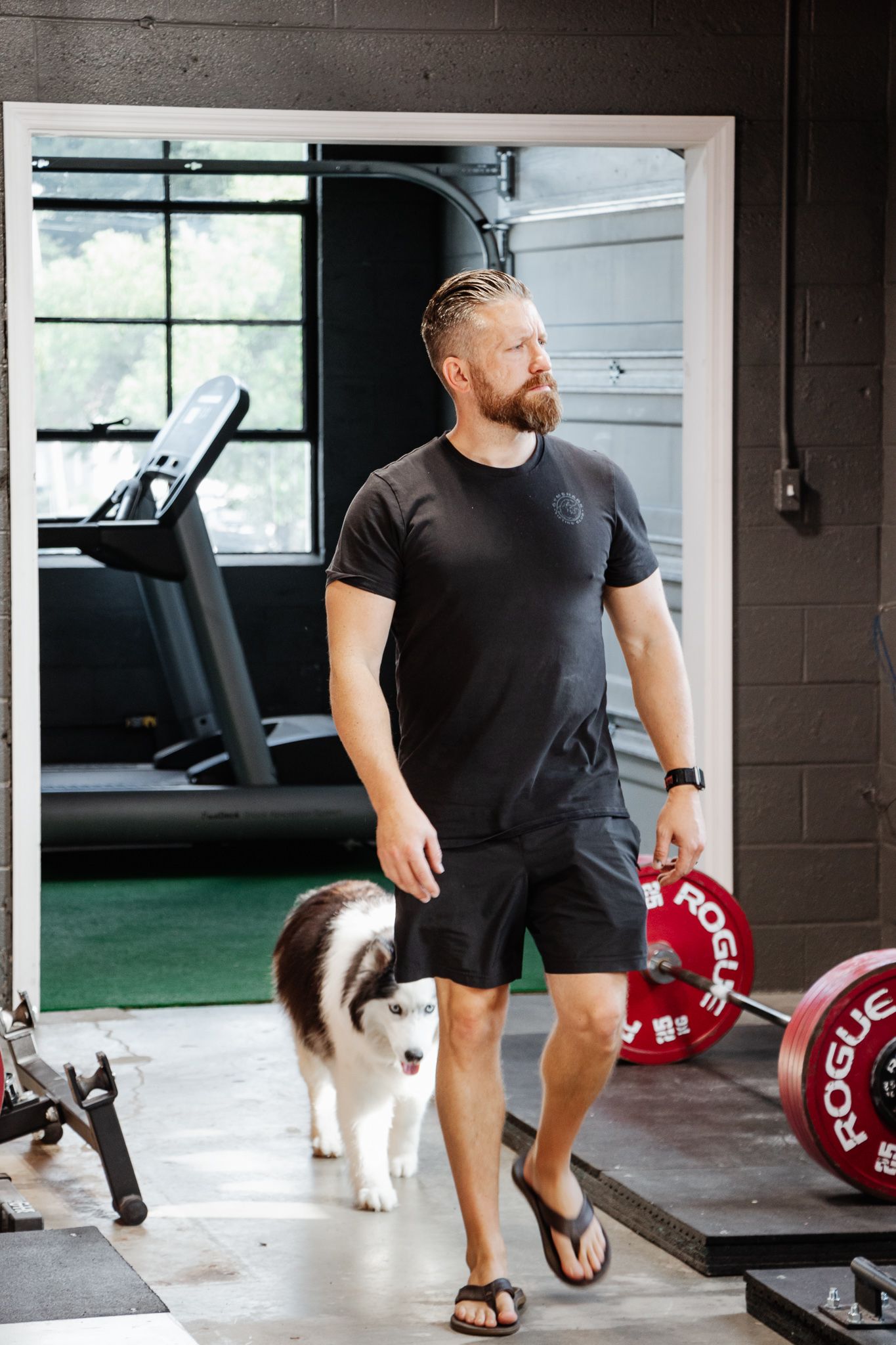 Man in black workout clothes walks in a gym with a husky following behind; weight plates and treadmill visible in the background