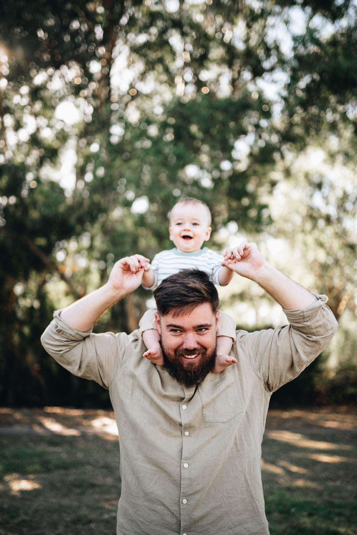 A father holds a baby on their shoulders in a sunny outdoor setting with trees in background taken by Melbourne family photographer.