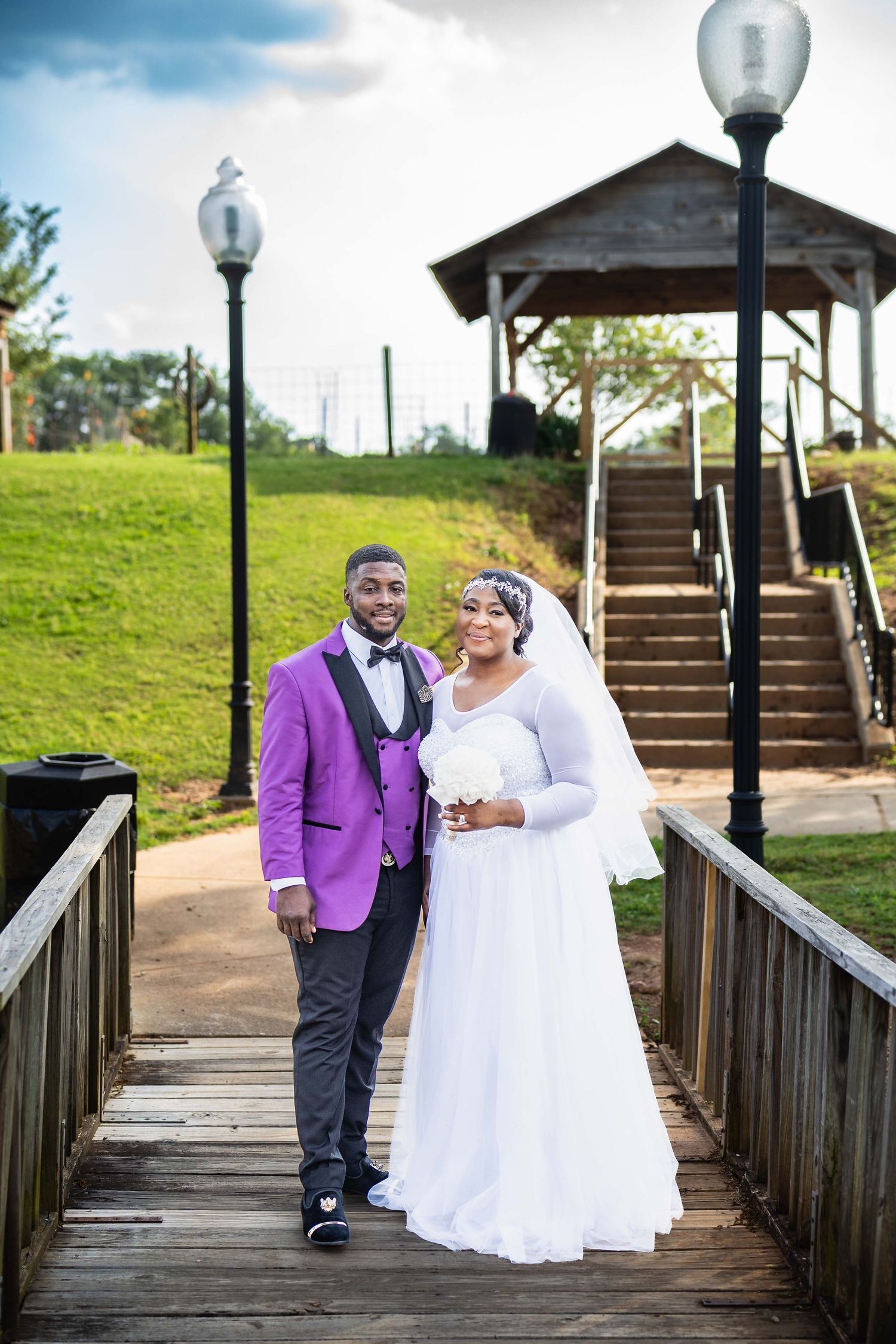 A couple in wedding attire poses on a wooden boardwalk with lamp posts and stairs in the background.