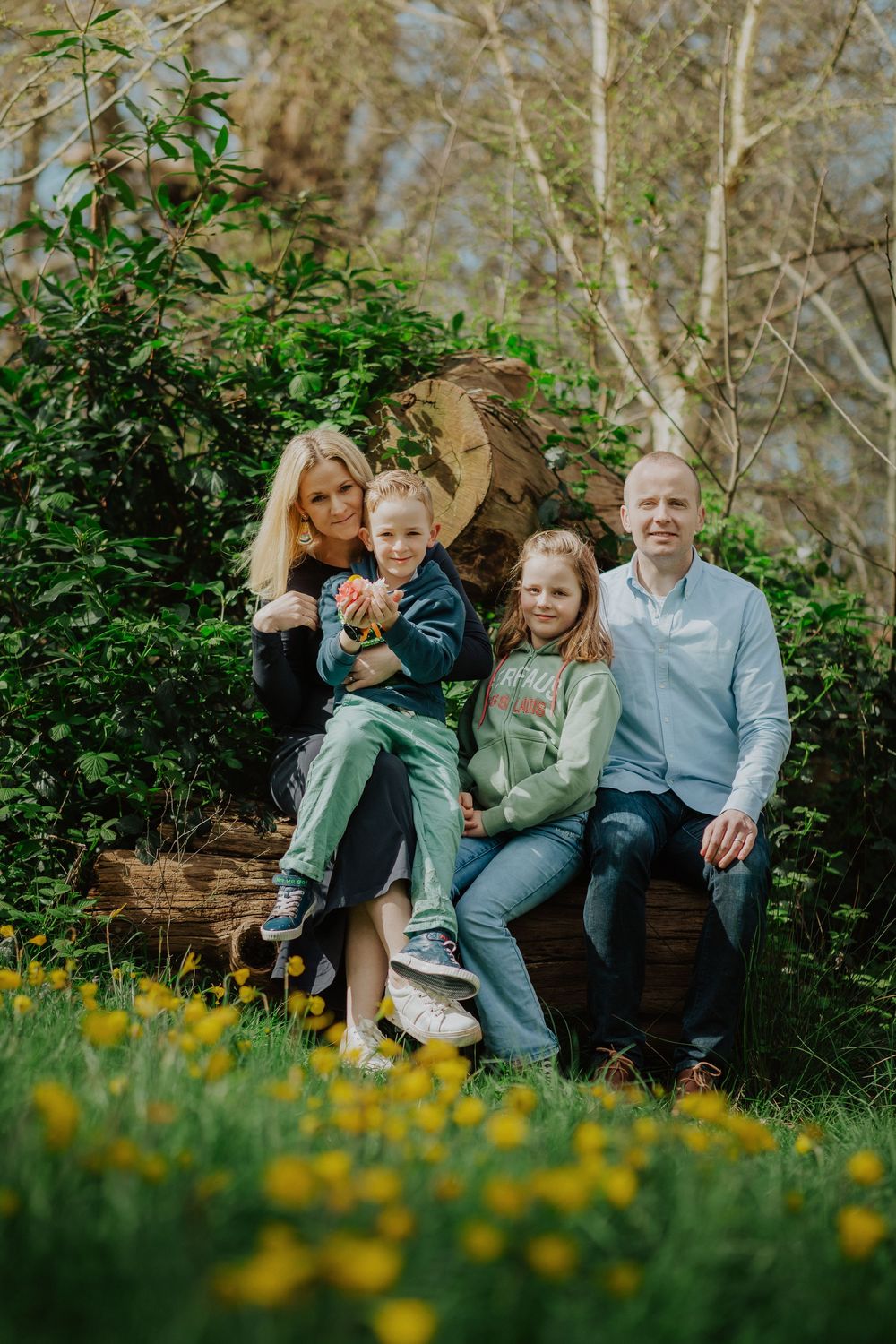 A family sits together on a log surrounded by green foliage and yellow wildflowers in an outdoor spring setting.