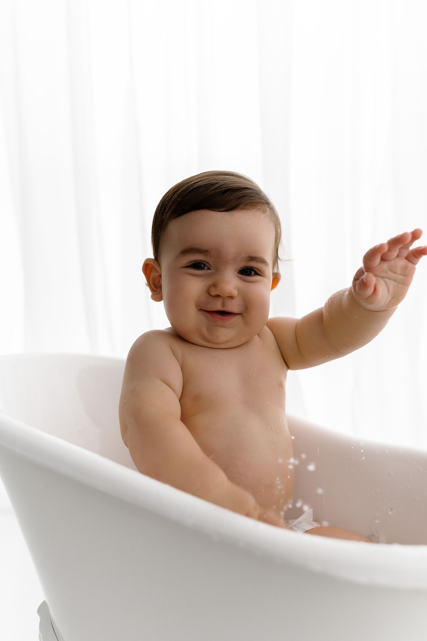 A cheerful baby splashes and waves during bath time in a white bathtub against a bright background.