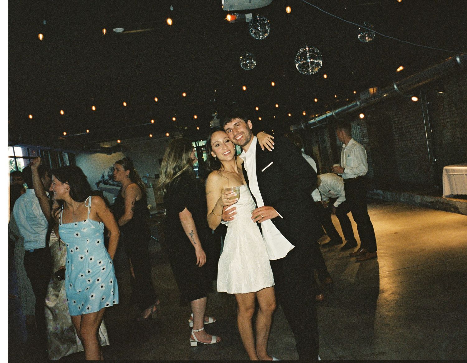 Couples dancing at a dimly lit party under string lights and disco balls.