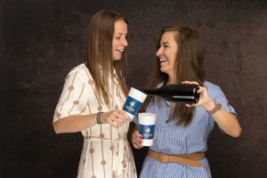 Two friends in casual dresses laugh while pouring coffee into reusable travel mugs against a dark background.