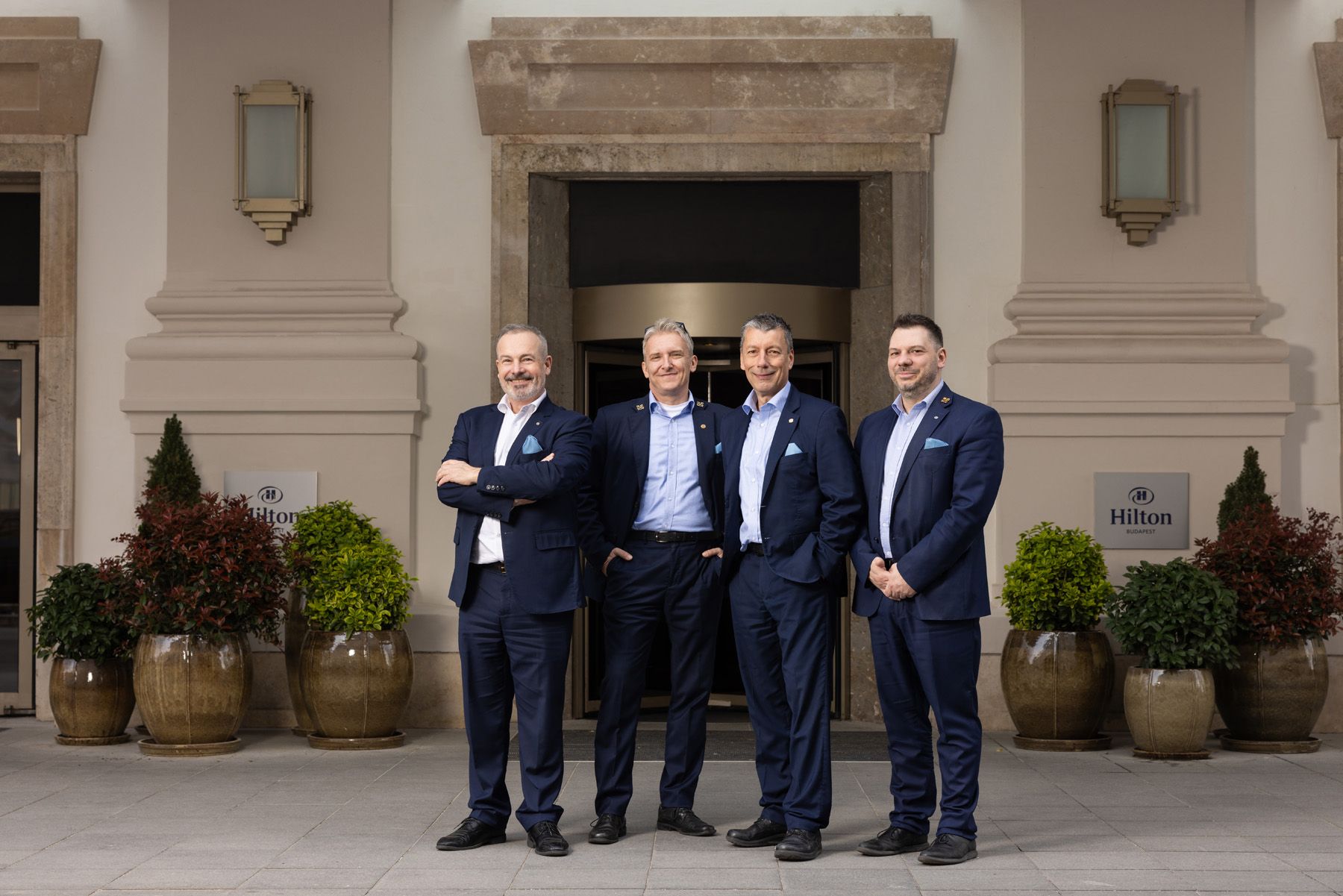 A line of people wearing navy blue suits stand against a light colored building exterior with decorative urns.