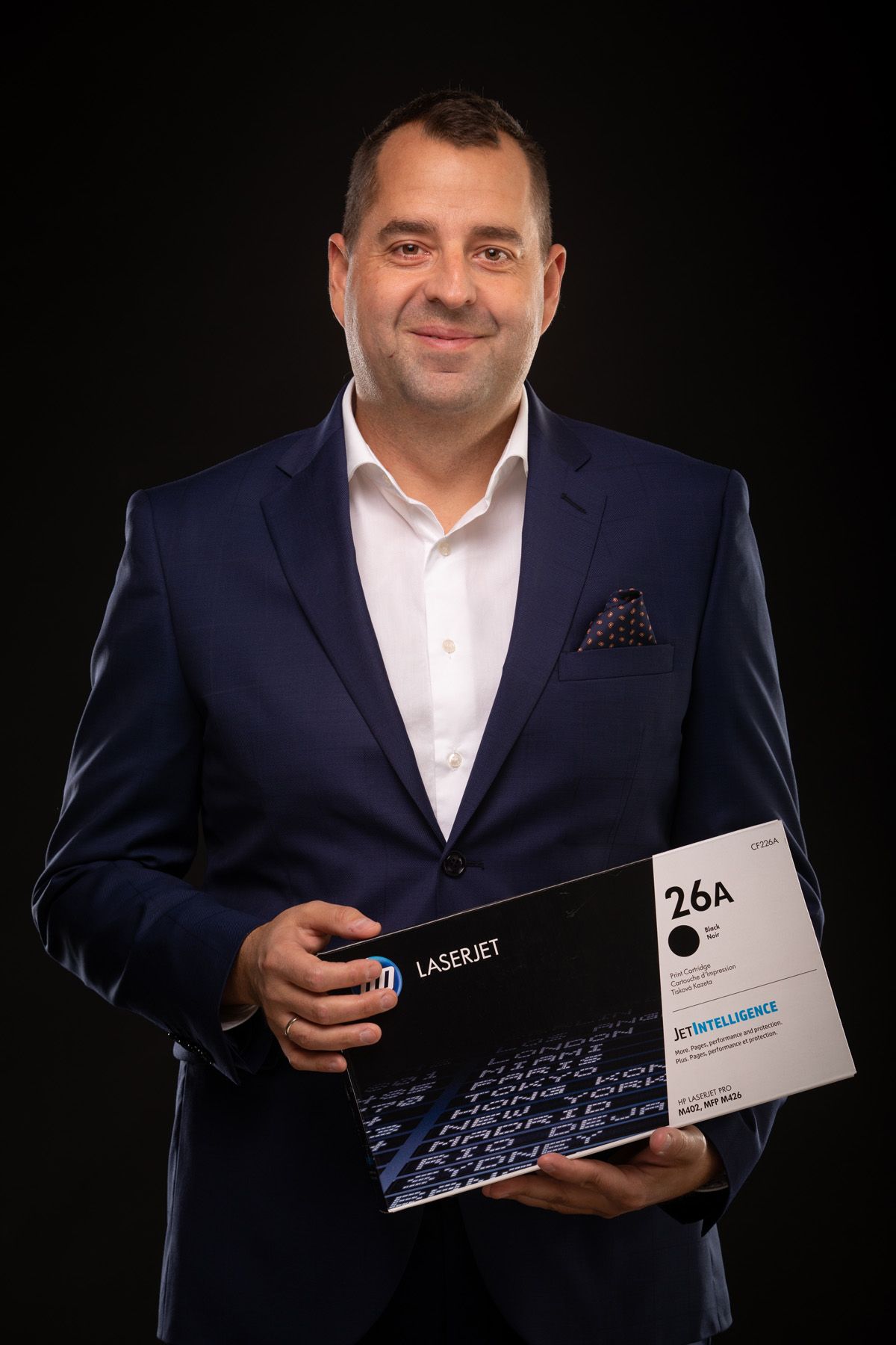 Professional portrait of someone in a navy suit holding business documents and materials against a black background.