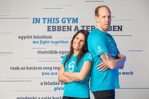 Two fitness instructors in matching turquoise shirts pose against a wall with motivational text at a gym.