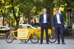 Two professionals in business suits stand in front of a yellow cargo delivery bike parked on a tree-lined street.