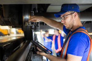 A worker in a blue uniform and glasses uses a digital tablet while inspecting equipment in an industrial setting.