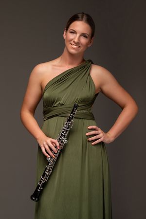 Professional musician in an elegant olive green one-shoulder gown holding an oboe against a dark background.