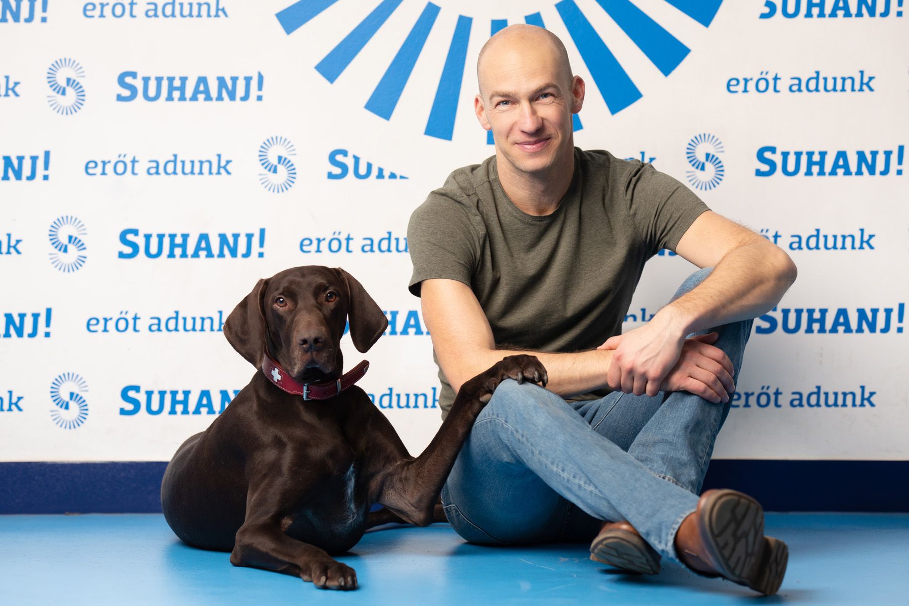 Casual portrait of someone sitting next to a chocolate labrador puppy against a branded backdrop with repeating blue logos.