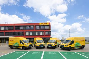 Four yellow DHL delivery vans parked in front of a red commercial building on green marked parking spots under blue sky.