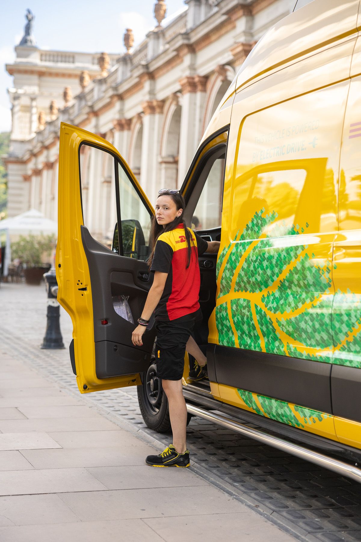 Delivery worker standing by yellow van with nature-themed decal parked next to ornate classical building.
