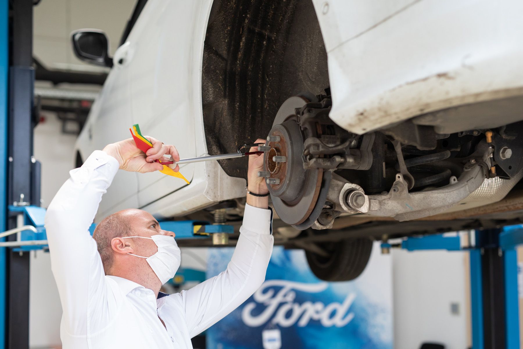 Auto mechanic in protective mask working underneath a vehicle at Ford service center.