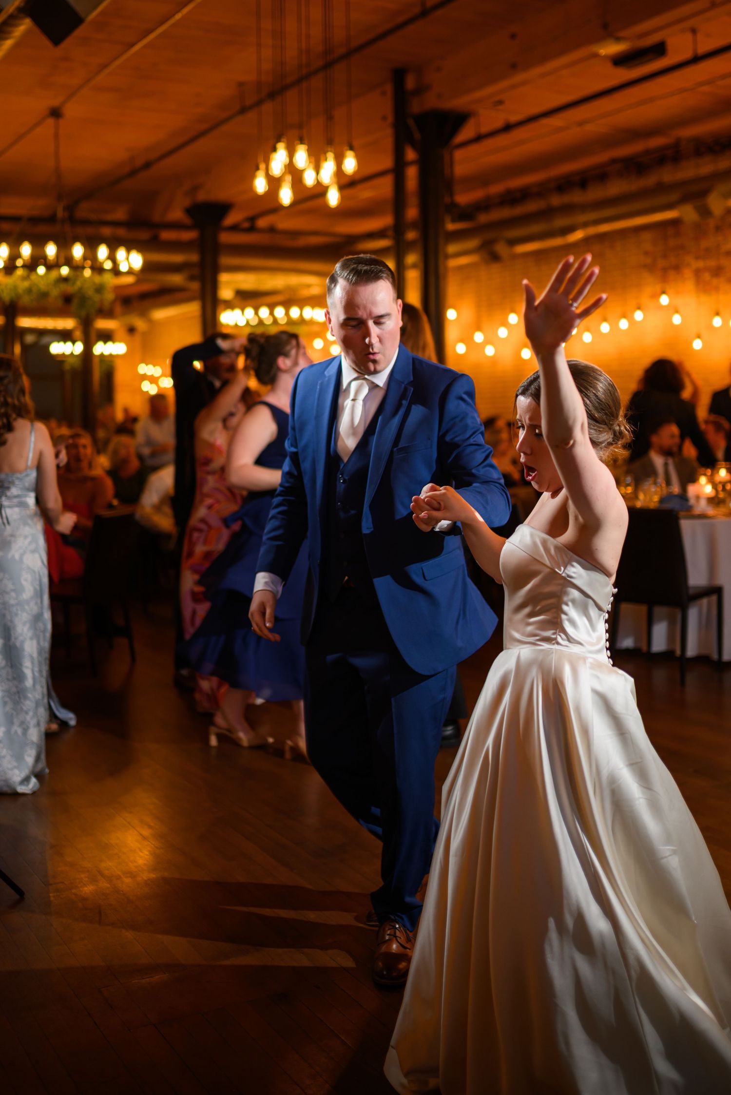 Couple dancing at a wedding reception in a warmly lit venue with chandeliers and a festive atmosphere.