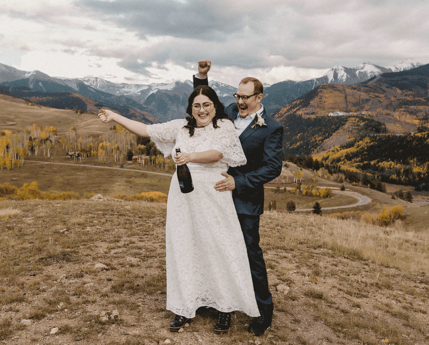 A couple in wedding attire poses joyfully on a hilltop overlooking autumn mountains in Colorado.