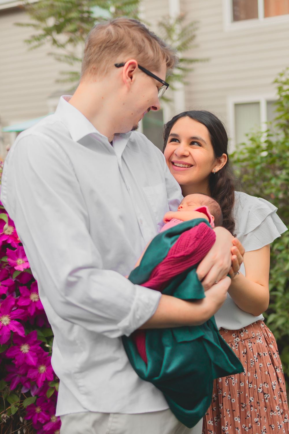 A couple shares a loving moment outside while holding a baby wrapped in a green and pink blanket near vibrant magenta flowers.