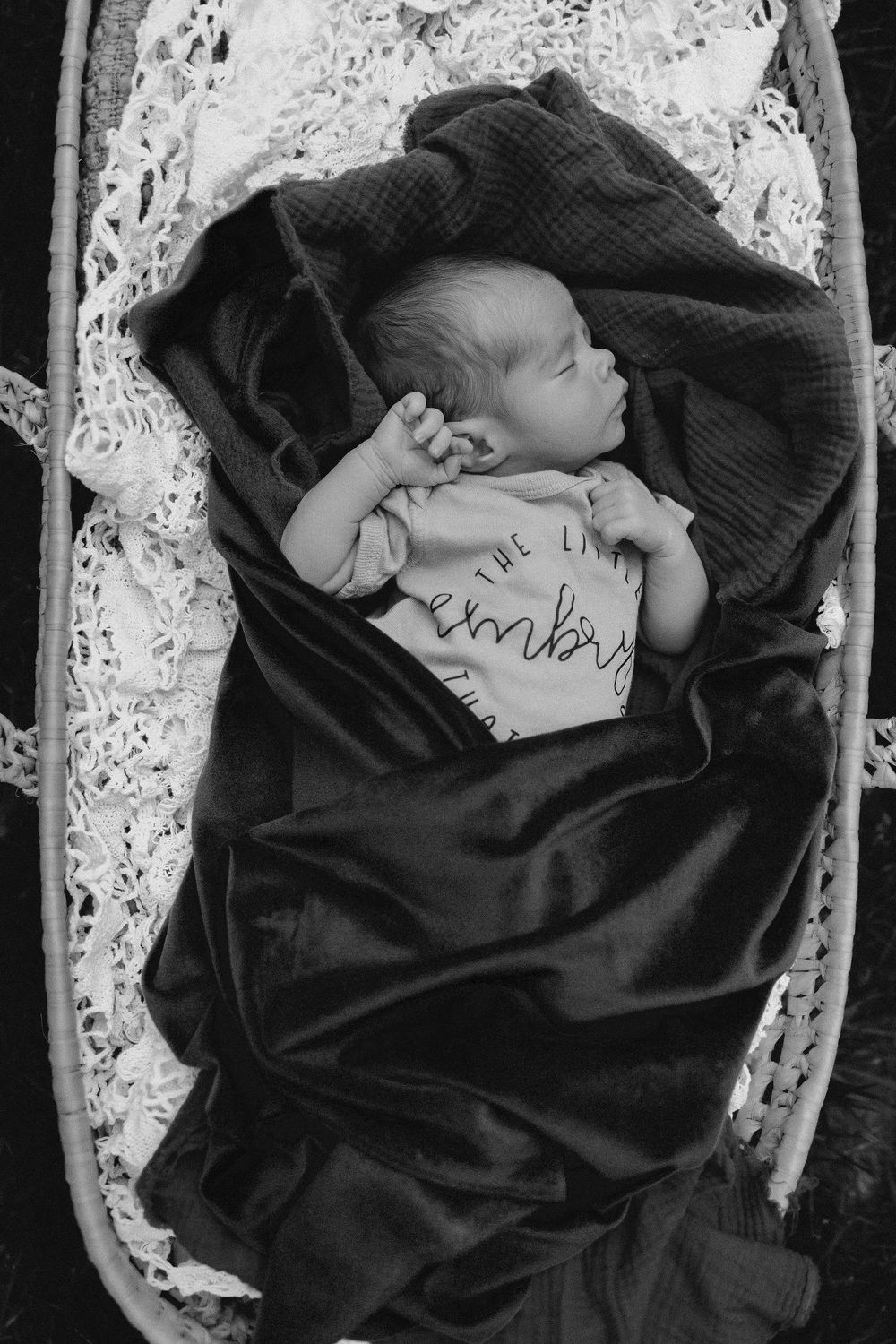 A sleeping baby wrapped in a dark blanket lies in a lace-lined bassinet, wearing a onesie with text.  Black and white image