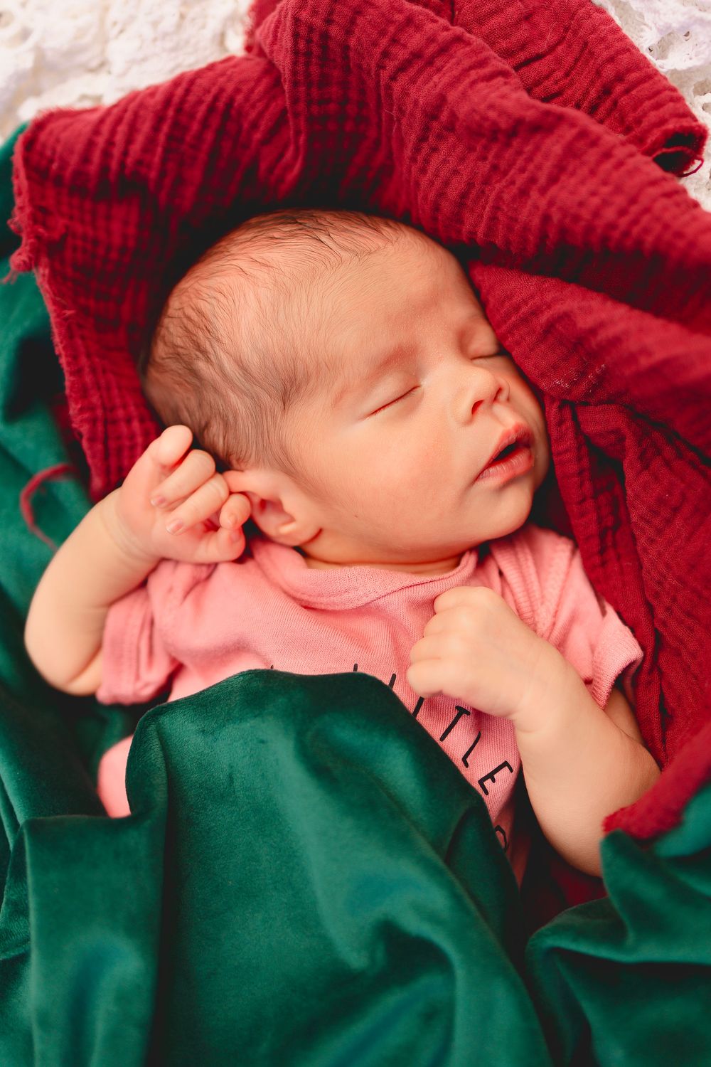 Peaceful sleeping newborn wrapped in red and green blankets wearing a pink outfit.