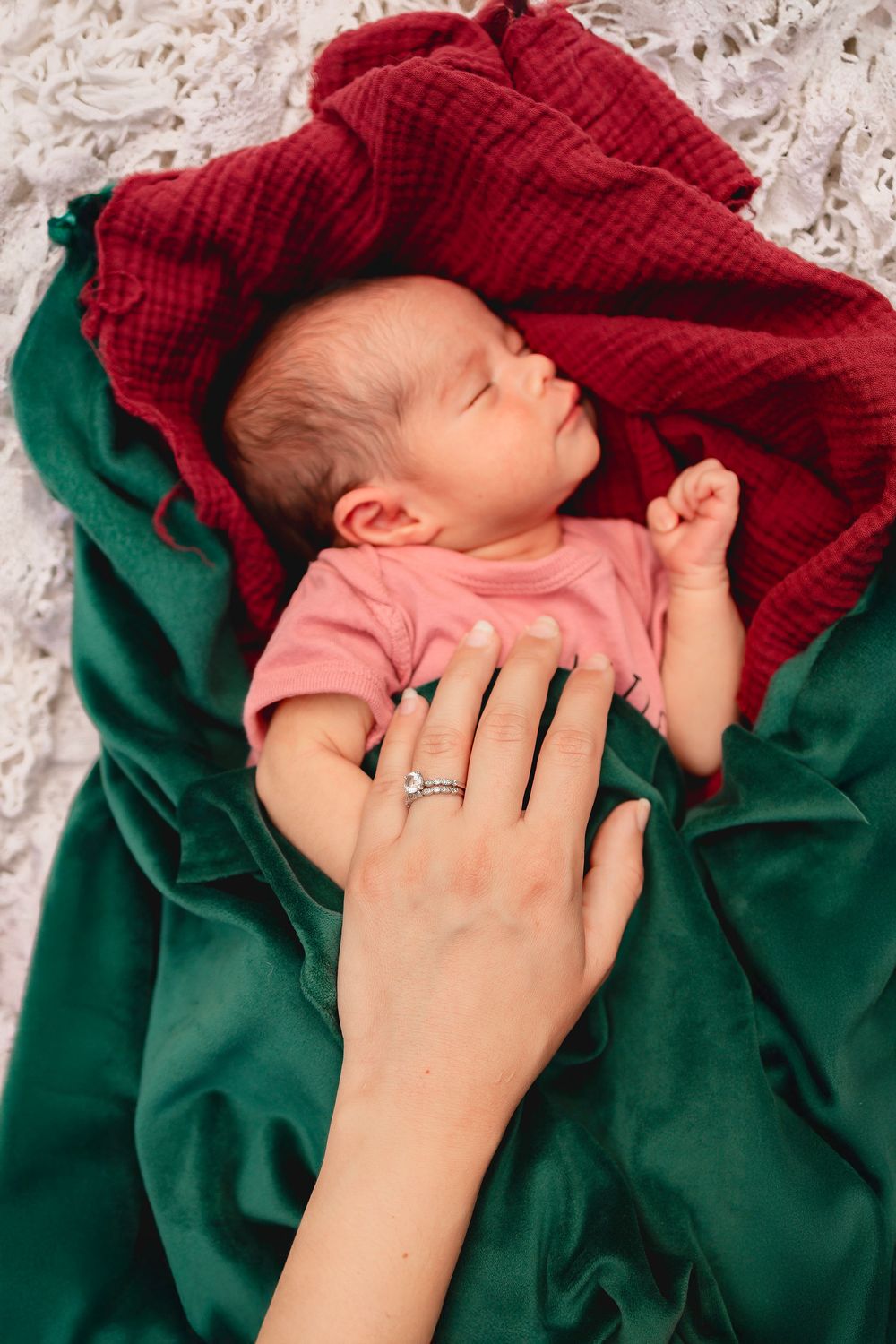 A newborn baby sleeping peacefully wrapped in cozy burgundy and emerald green blankets while a hand gently rests on baby