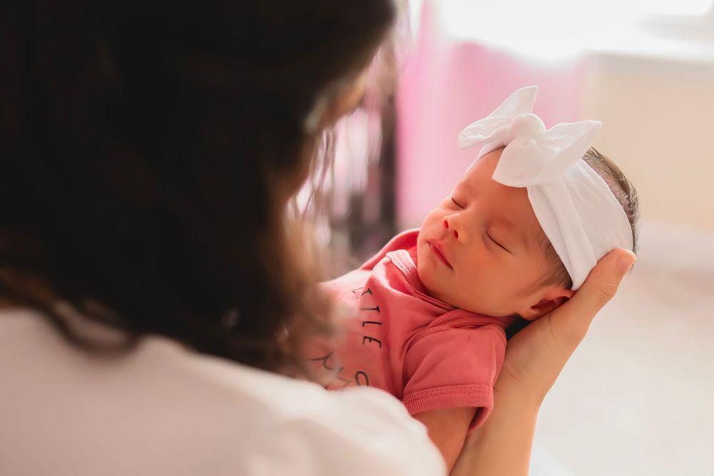 Newborn in a white bow headband sleeping peacefully in soft lighting.  Mother holding her