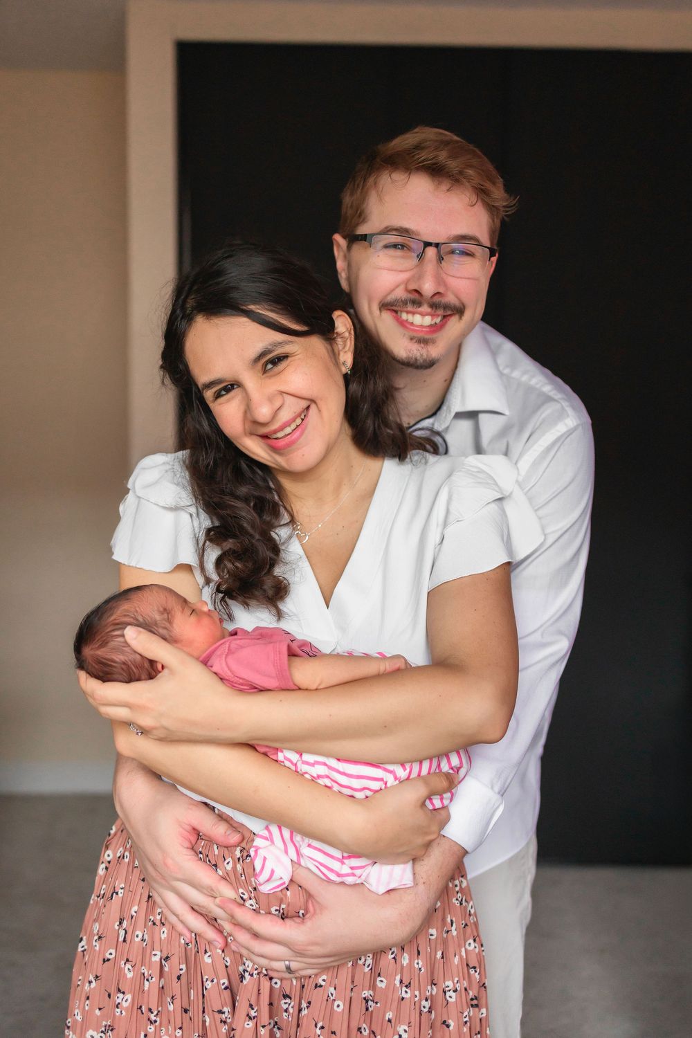 A couple embraces and smiles together.  Mom holding newborn baby girl