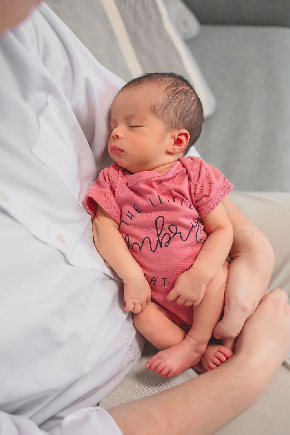 A sleeping newborn baby wearing a pink onesie rests peacefully in their caregiver's lap.