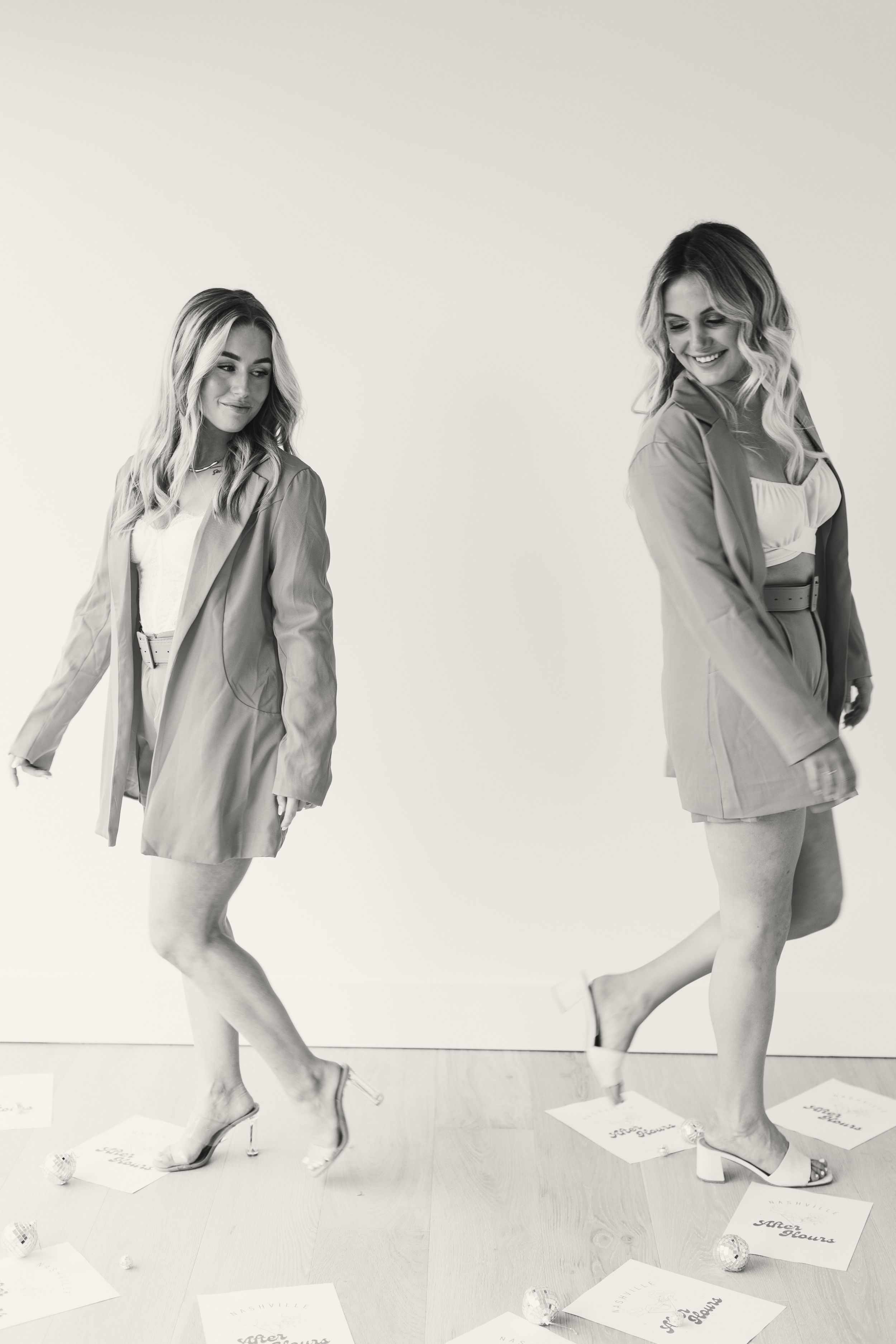 Two people in light-colored outfits dancing and posing against a white wall in a casual black and white photoshoot.