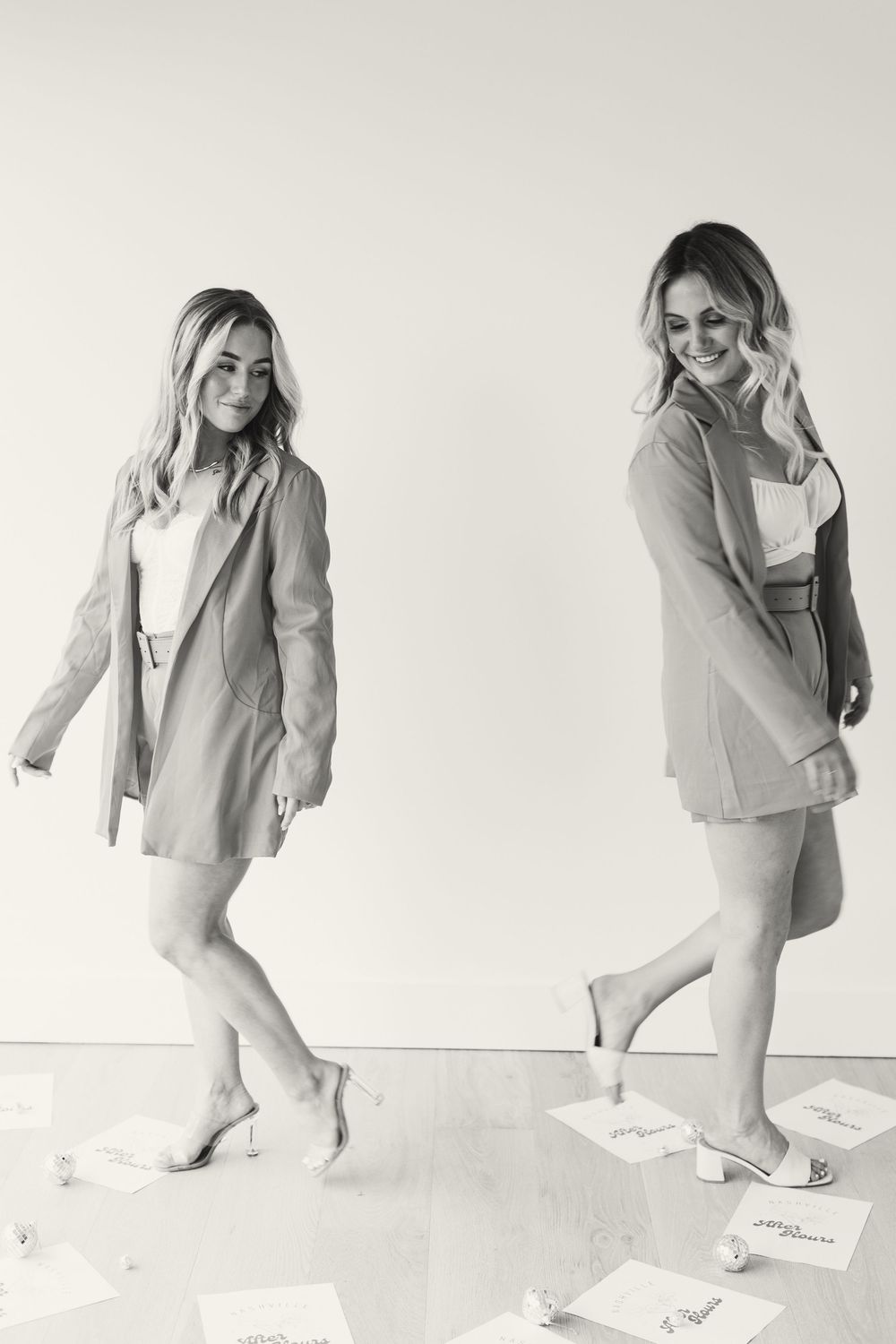 Two people in light-colored outfits dancing and posing against a white wall in a casual black and white photoshoot.