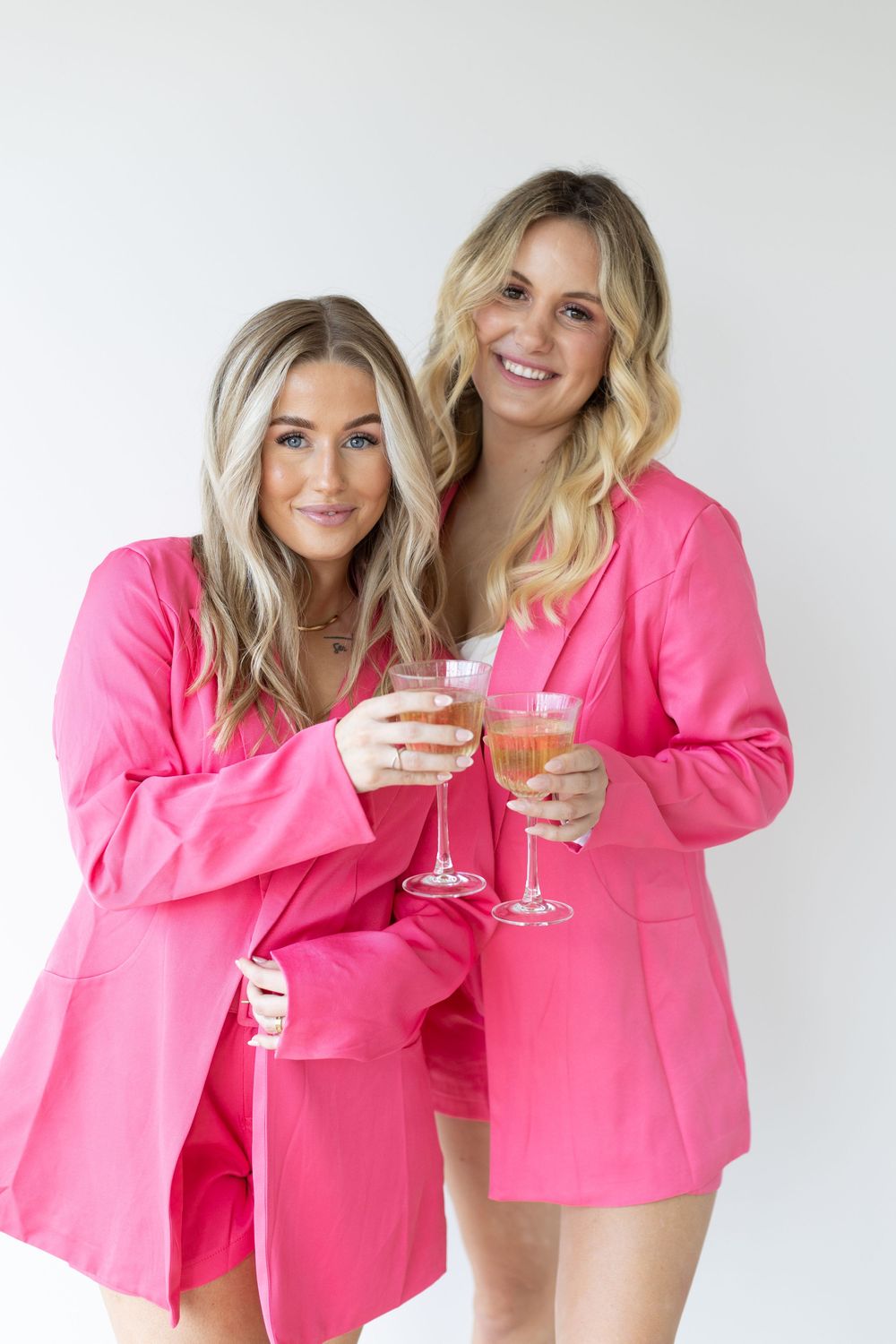 Two people in bright pink blazers holding champagne glasses pose against white background in a fashion photoshoot.