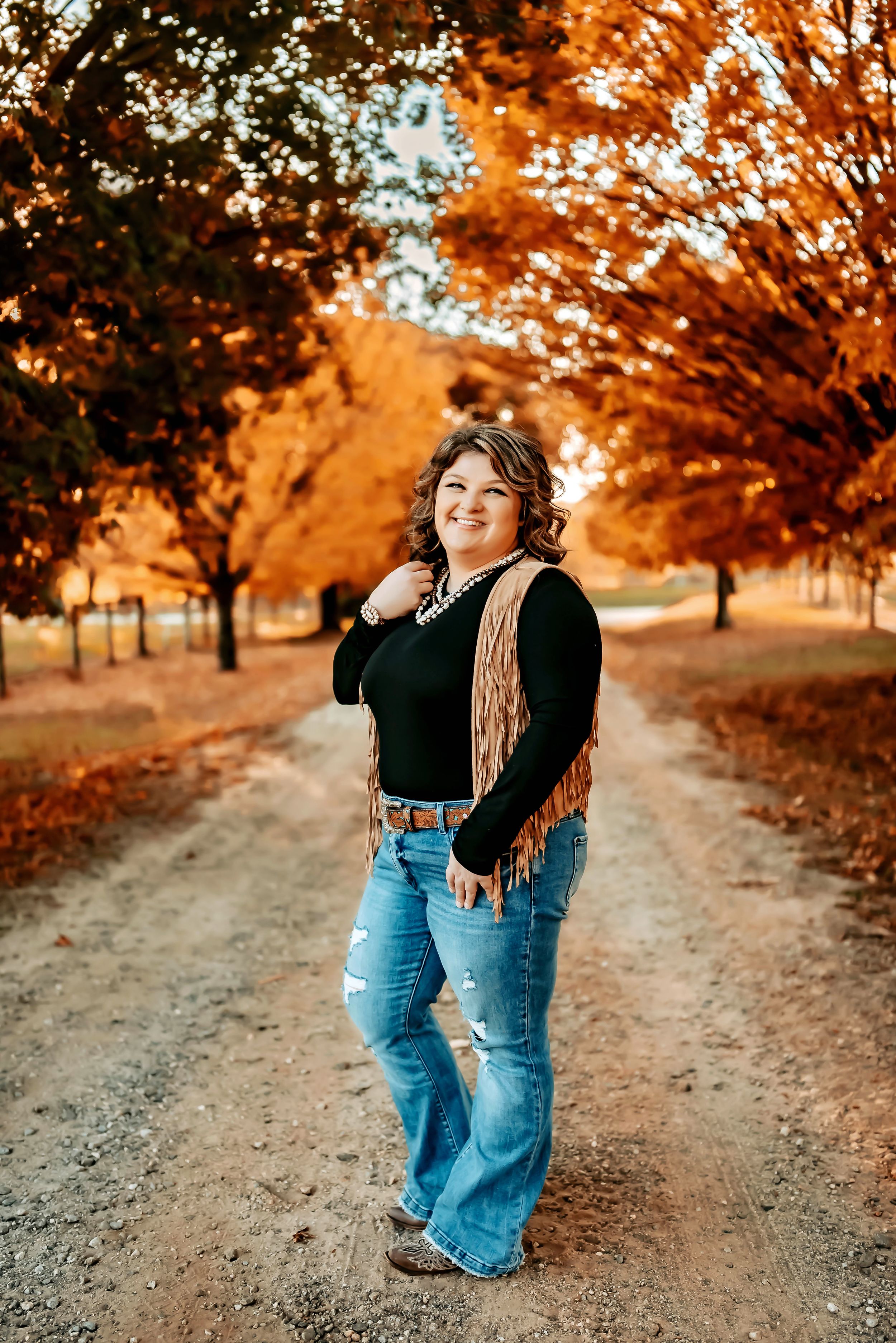 Person in casual fall outfit standing on dirt path with orange autumn foliage in background.