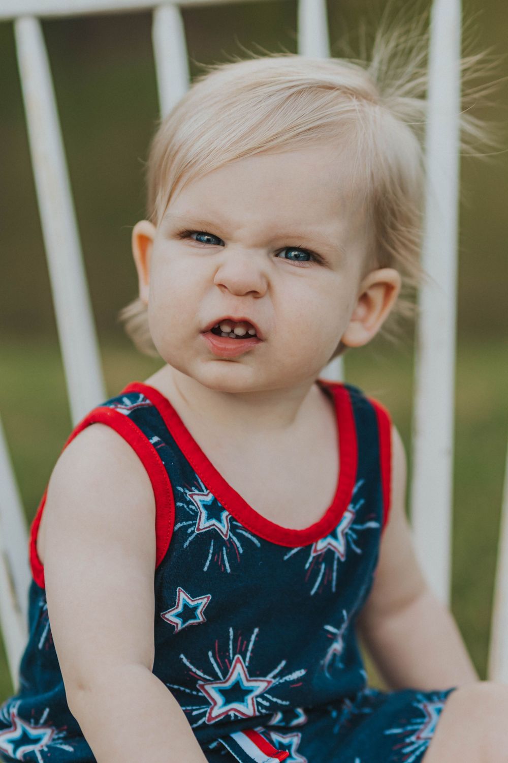 A toddler wearing a blue and red star-patterned tank top makes an expressive face while sitting outdoors.