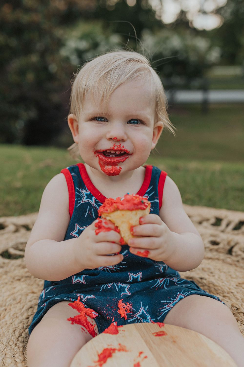 A messy-faced toddler enjoys eating watermelon while wearing a patriotic outfit outdoors.