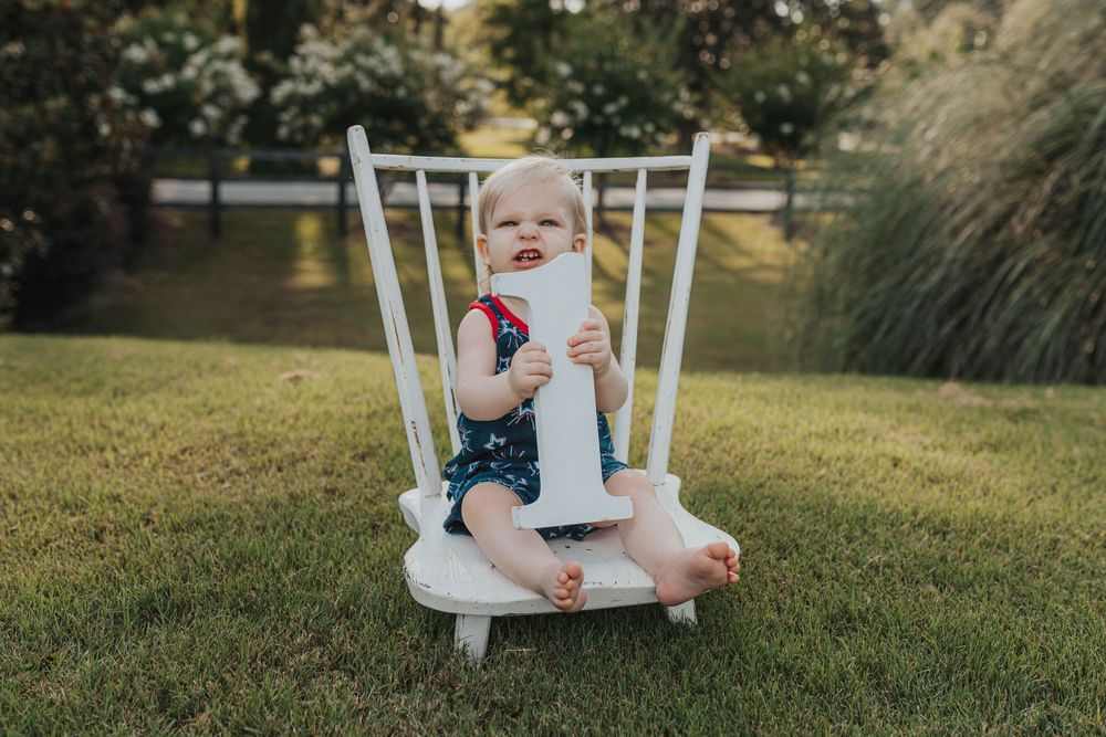 A toddler sits in a white wooden chair on a sunny lawn wearing patriotic clothing.
