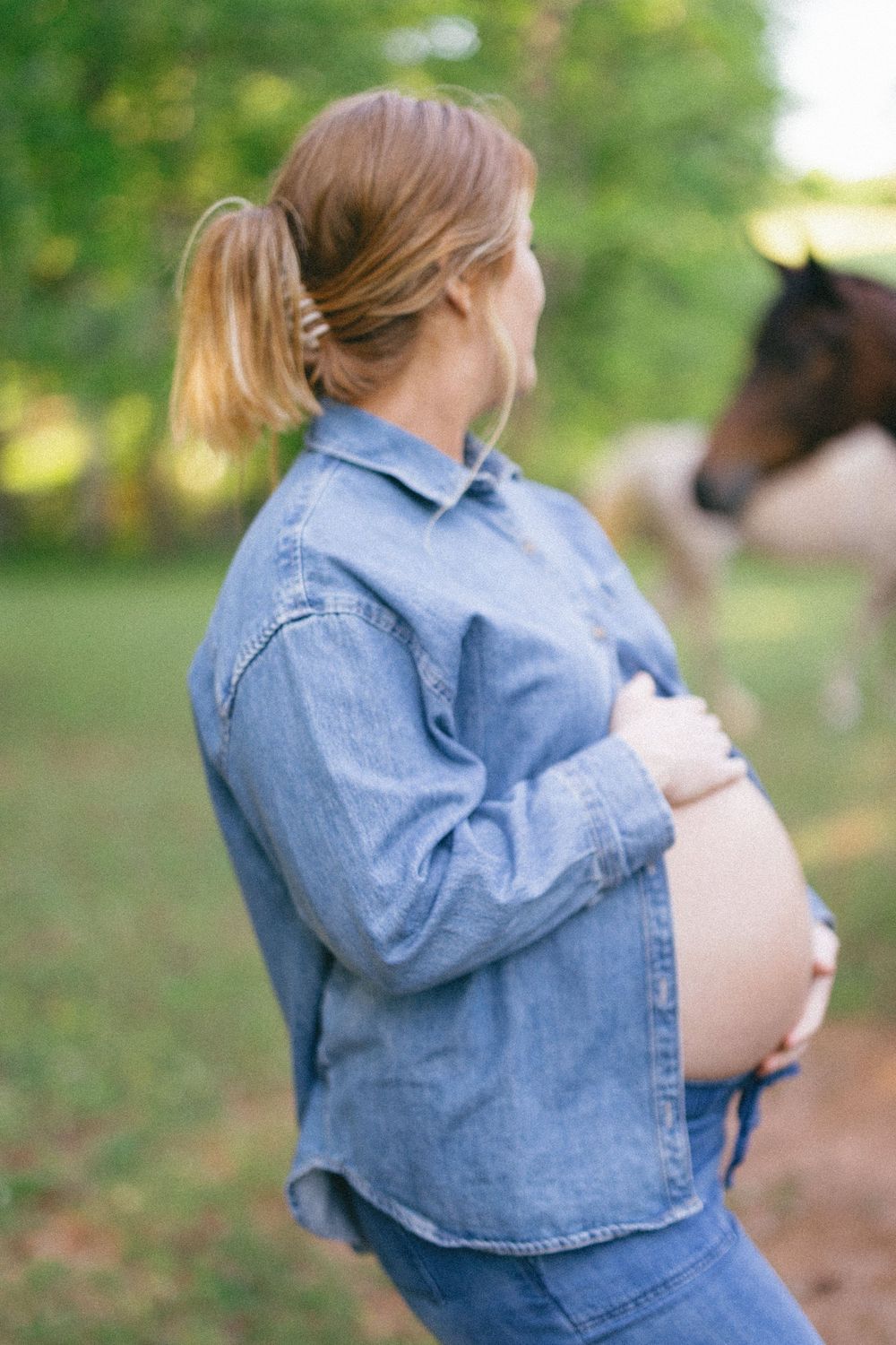 Expectant mother in denim outfit cradles her belly while standing outdoors with a horse visible in the background.