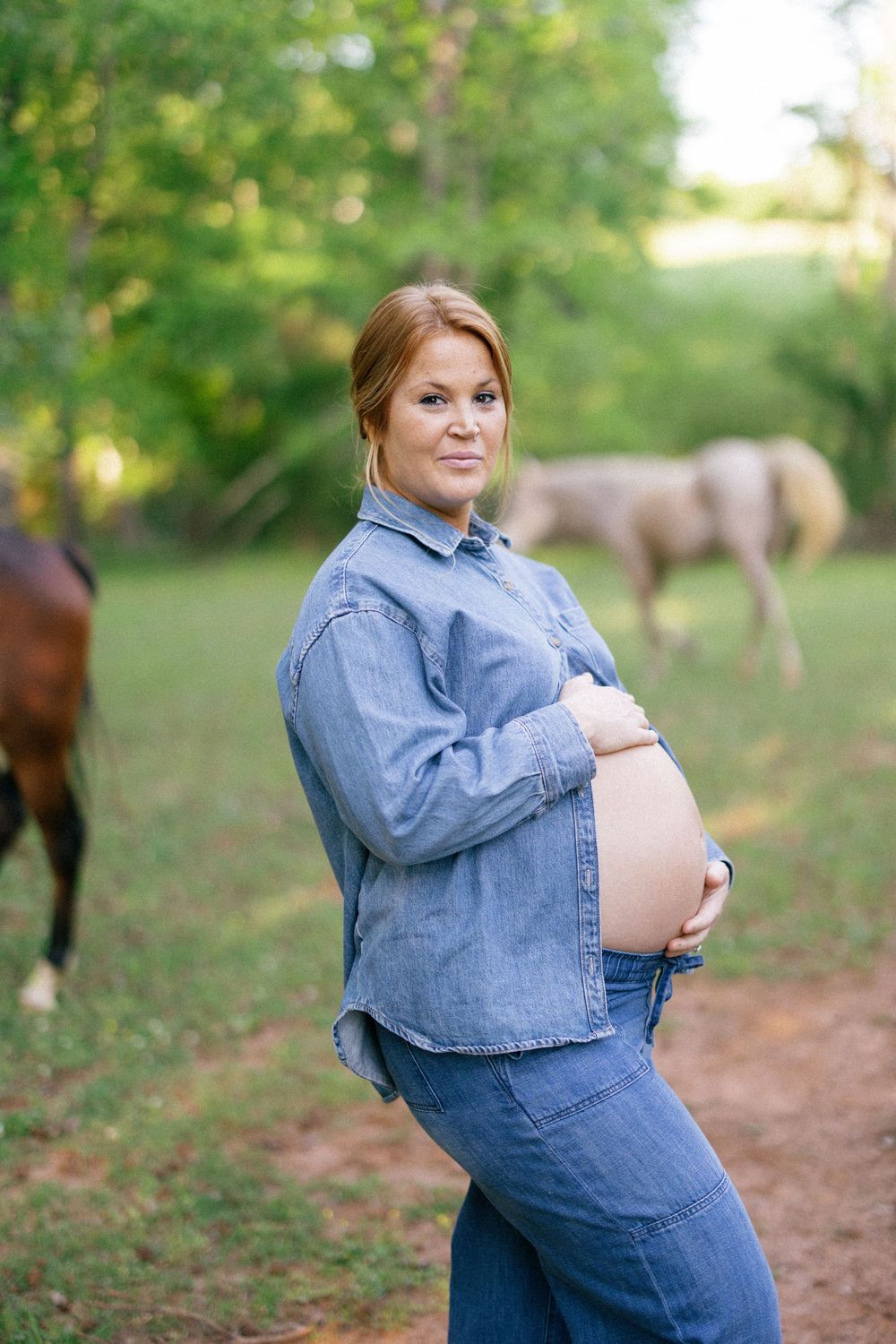 A pregnant person wearing denim stands in a grassy outdoor area with horses visible in the background.