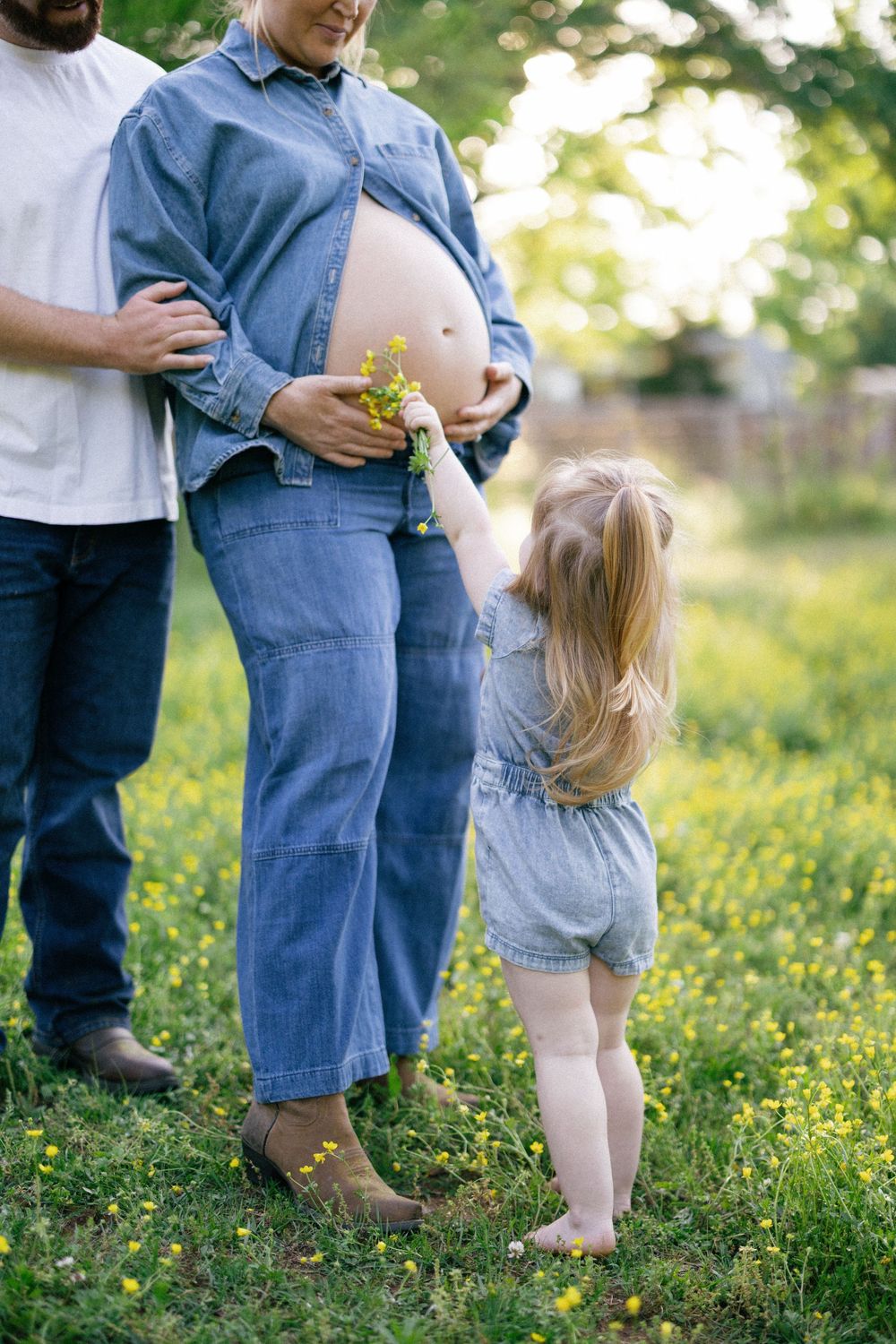 A small child in a denim dress touches a pregnant person's belly while standing in a sunlit garden with yellow flowers.
