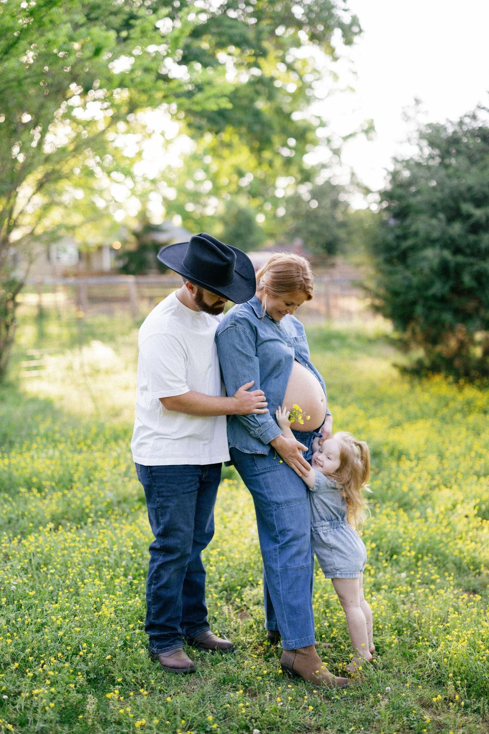 A family shares a tender moment together in a sunlit field with yellow wildflowers and green trees in the background.