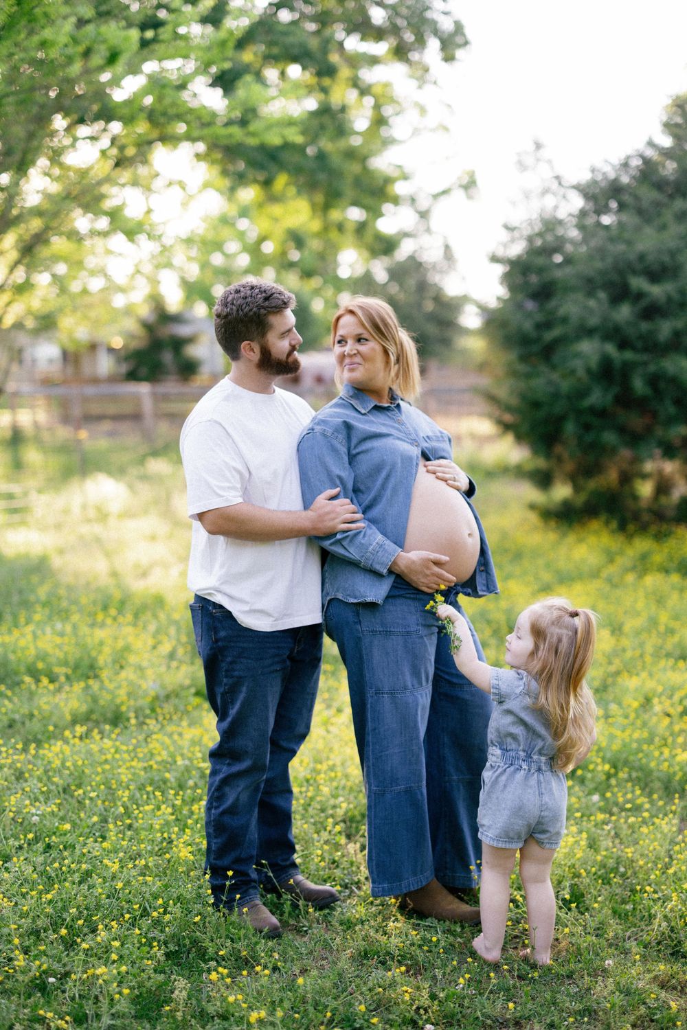 A family stands together outdoors in a sunlit garden with wildflowers as they await their new arrival.