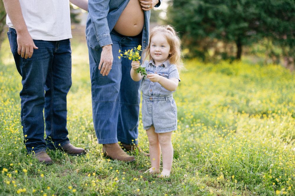 Someone in denim overalls stands with family members in a sunlit meadow filled with yellow wildflowers.