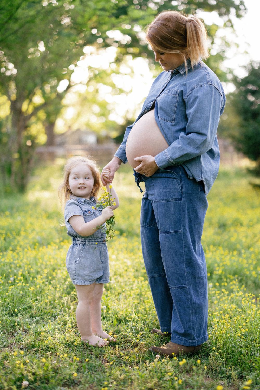 A pregnant woman in denim stands with a young child in a sunlit meadow filled with yellow wildflowers.