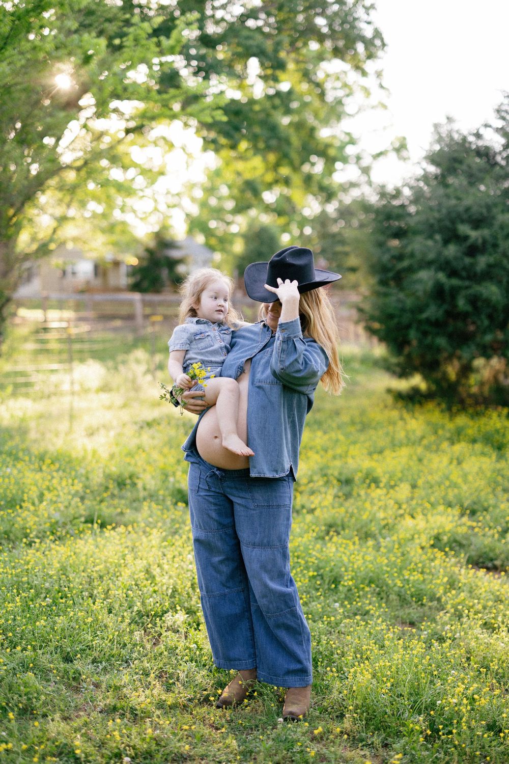 Child holding a bouquet plays with parents in a sunlit field of yellow wildflowers on a spring day.