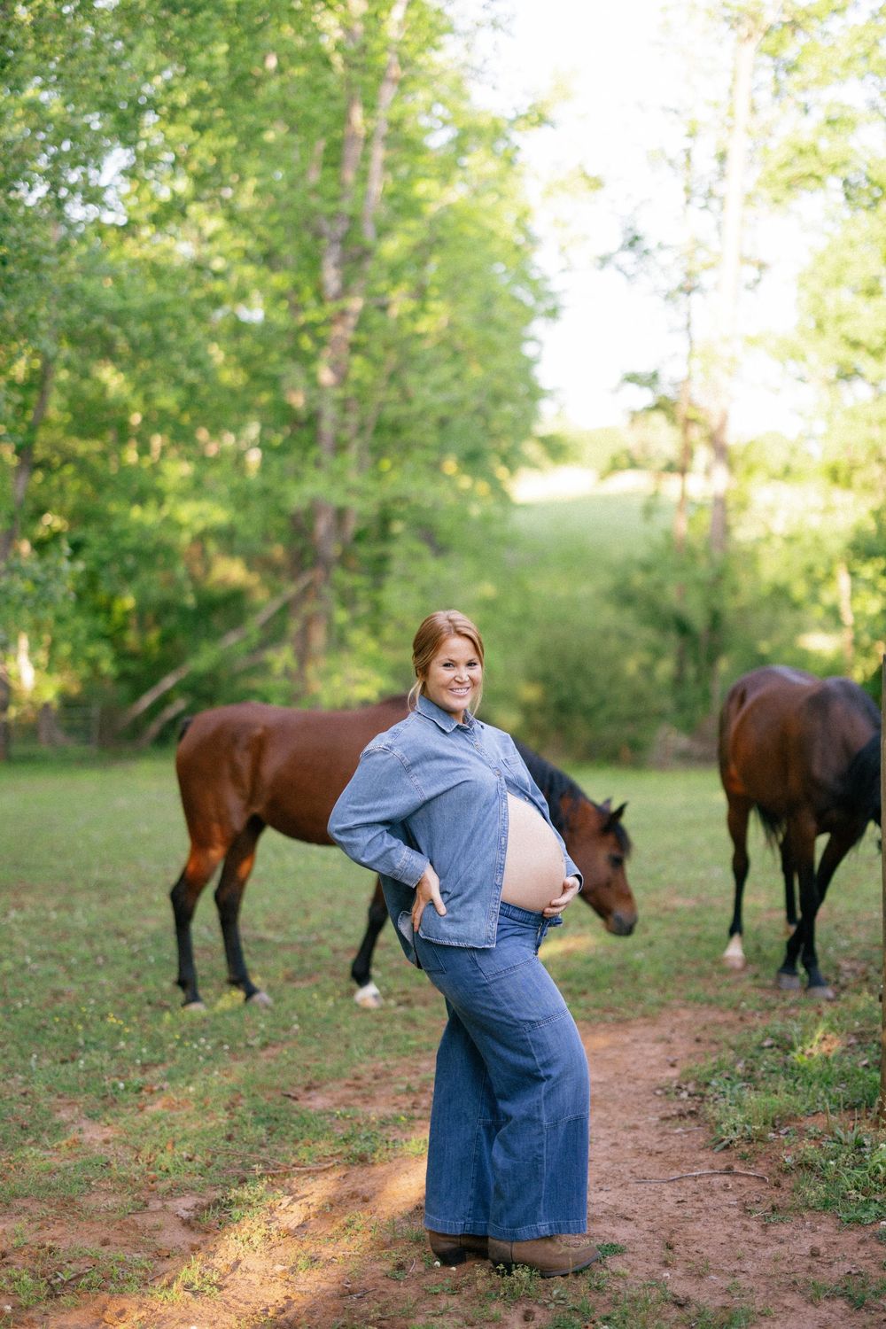 Pregnant woman in denim outfit stands on dirt path with two brown horses grazing in grassy field behind her.