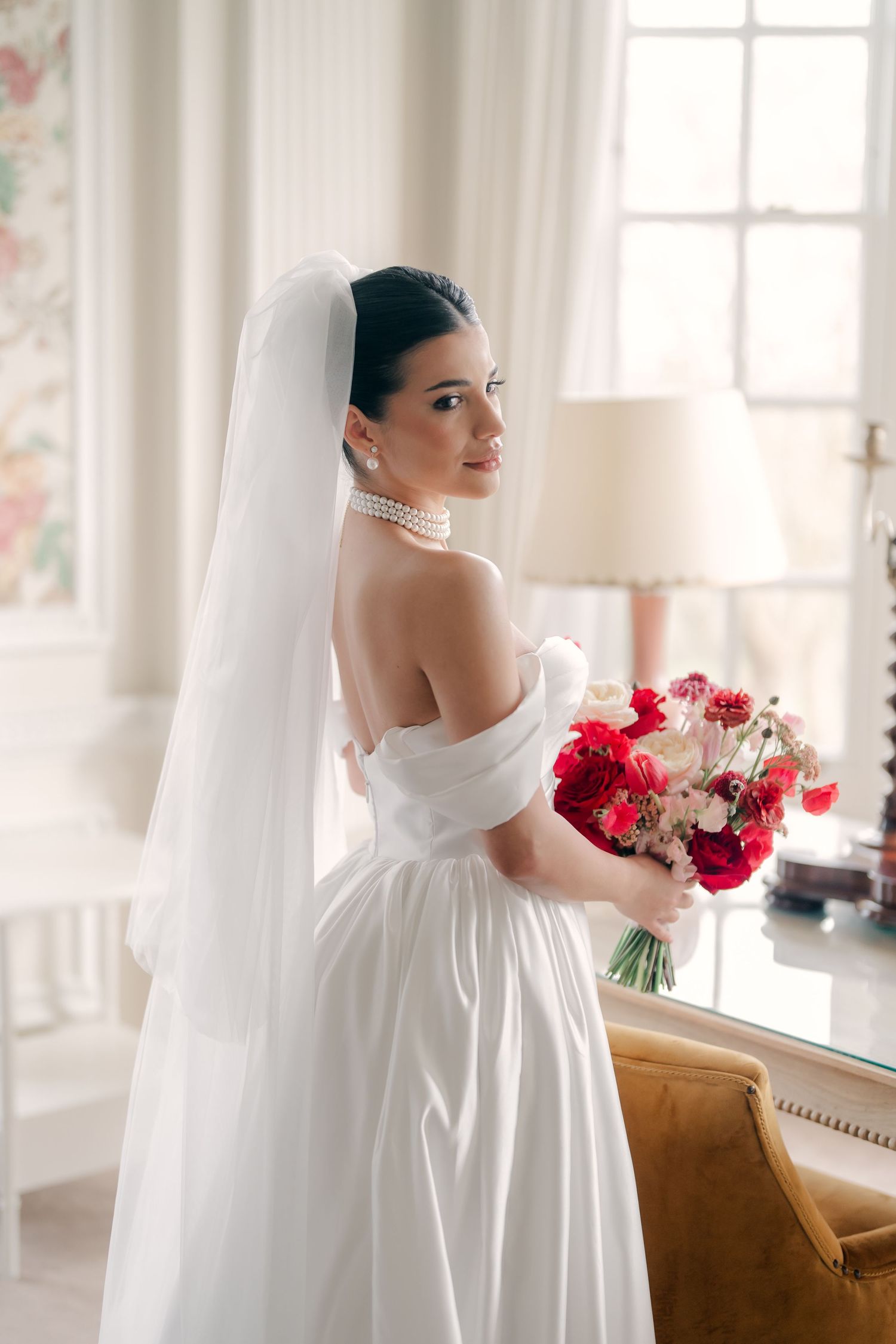 Romantic bridal portraits with red rose bouquet and pearl necklace in a light-filled room.