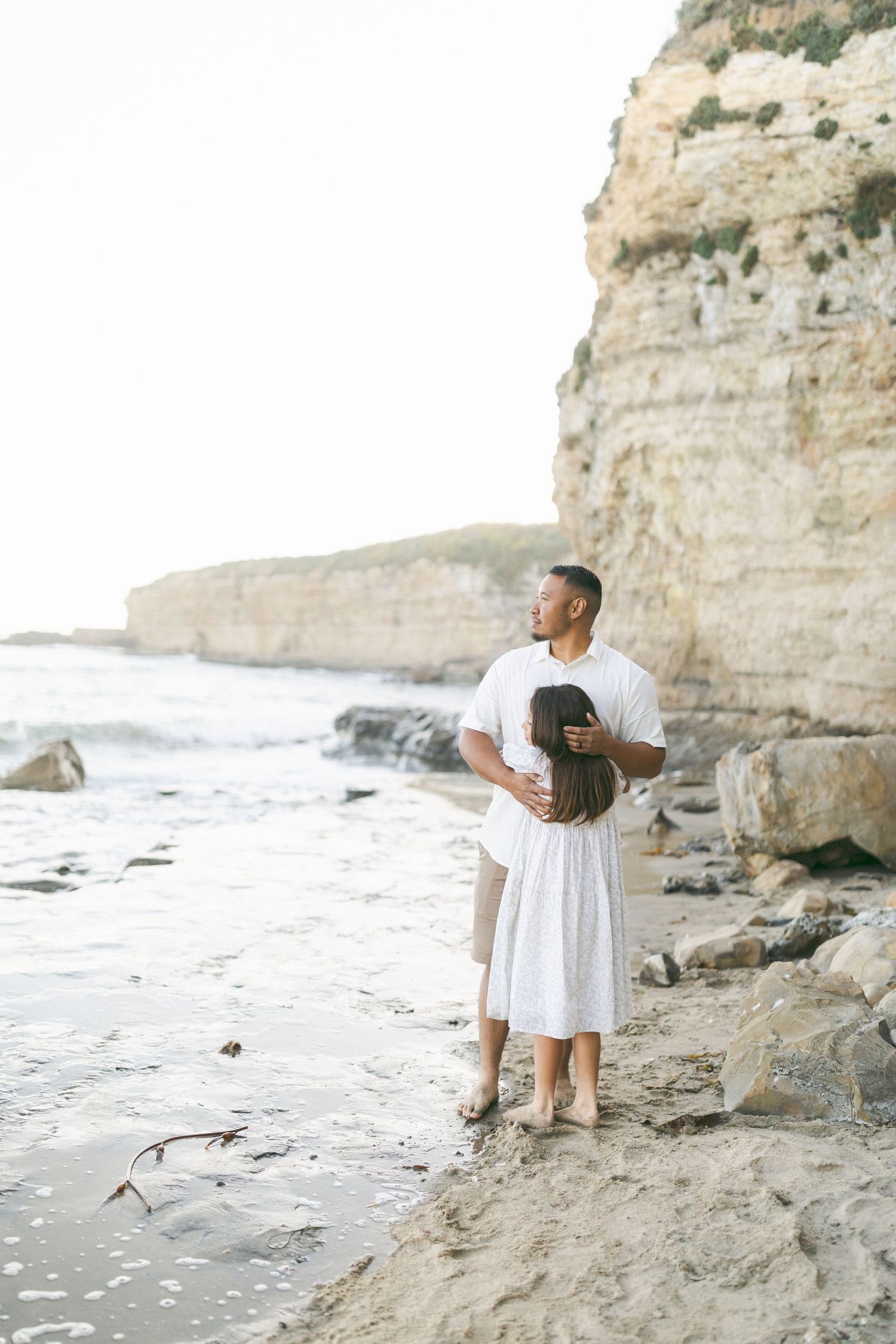 Couple in white outfits embrace while standing near ocean waves and dramatic coastal cliffs at sunset.