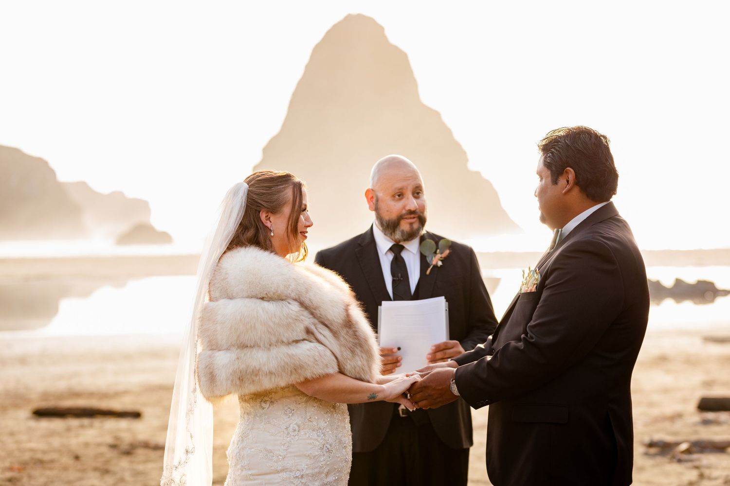 Bride and groom exchanging vows on sandy beach at golden sunset with dramatic sea stack rock formation behind.